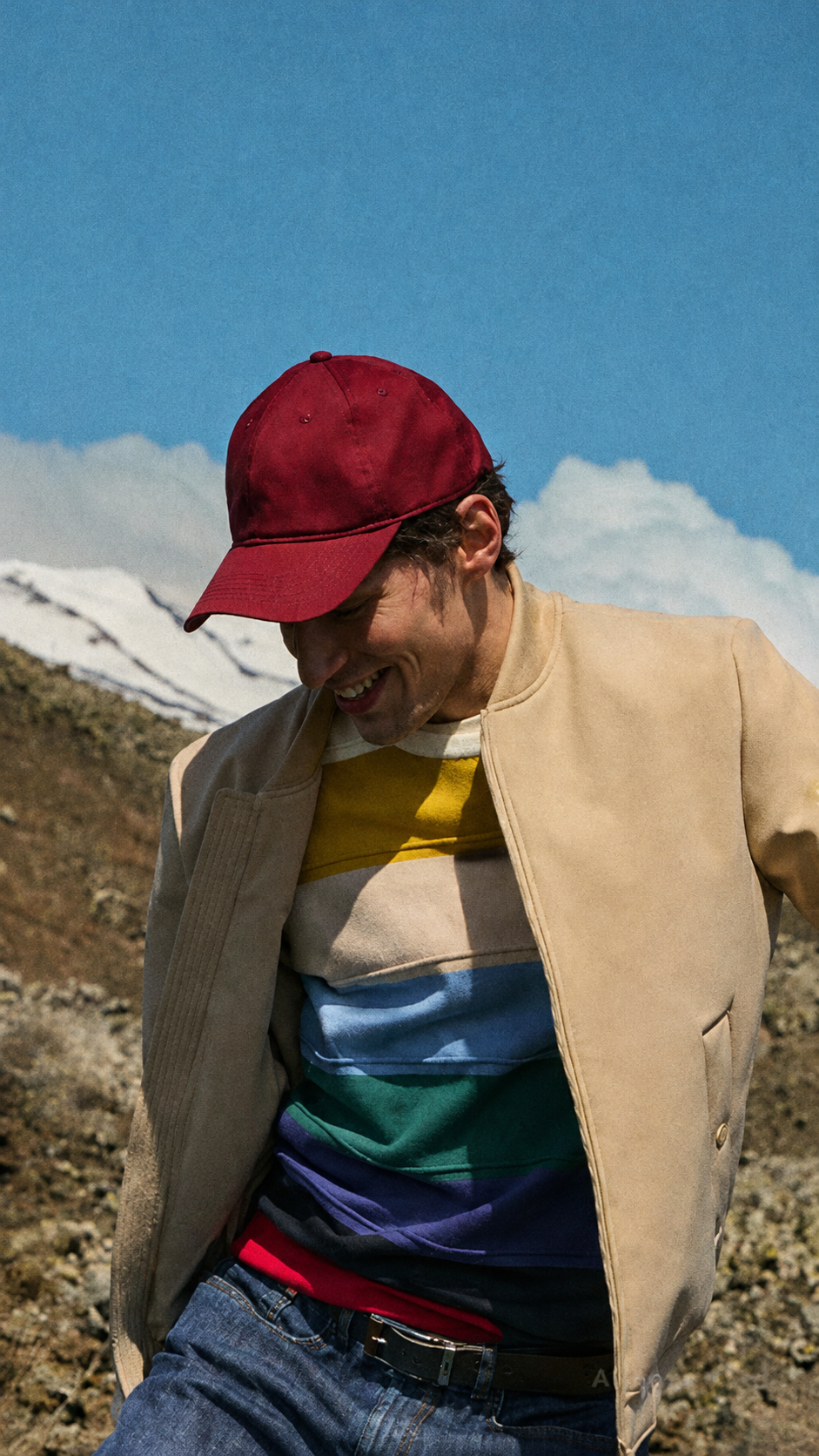 An image of a smiling man outdoors with a mountainous and volcanic landscape in the background. The man wears a burgundy baseball cap, an open beige bomber jacket, and a striped colorful sweater. He is looking downwards with an amused expression, in motion. The sky is blue with white clouds.