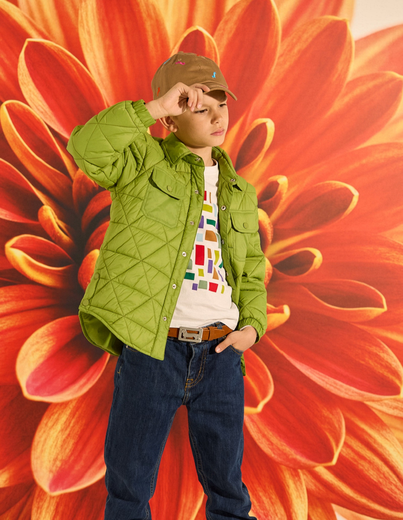 Bambino con giacca verde trapuntata, T-shirt stampata, jeans e cappellino beige