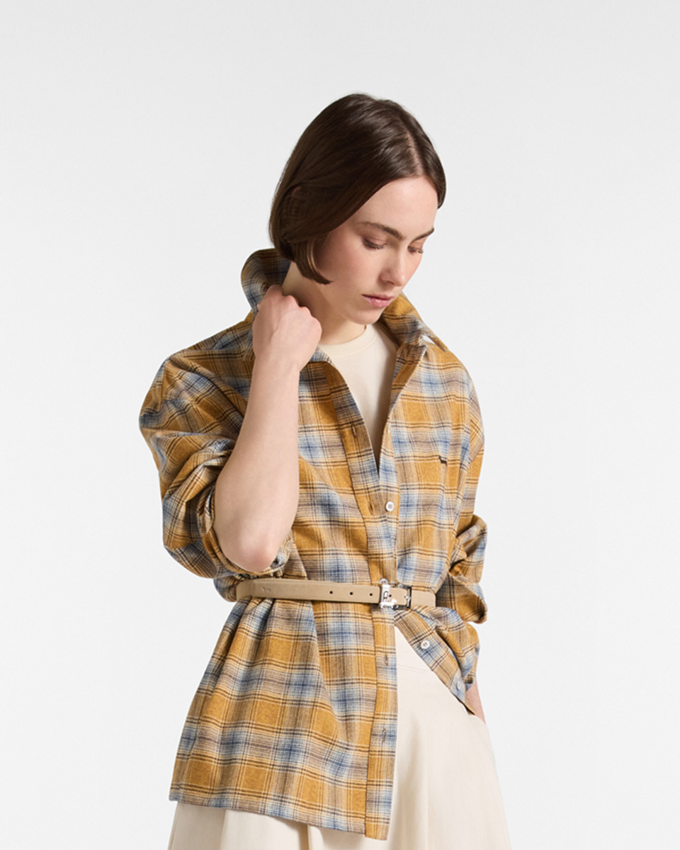 Female model sitting on a wire mesh chair, wearing an olive green checkered shirt with contrasting beige collar and cuffs, paired with dark jeans.