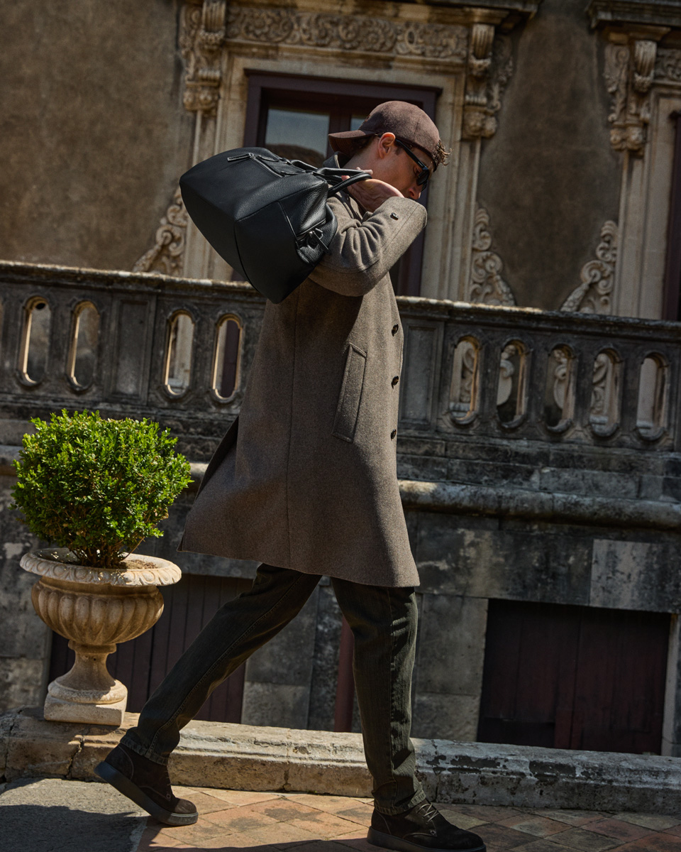 Male model in a grey coat, baseball cap, and sunglasses walking along a historic terrace while carrying a black leather travel bag over his shoulder.
