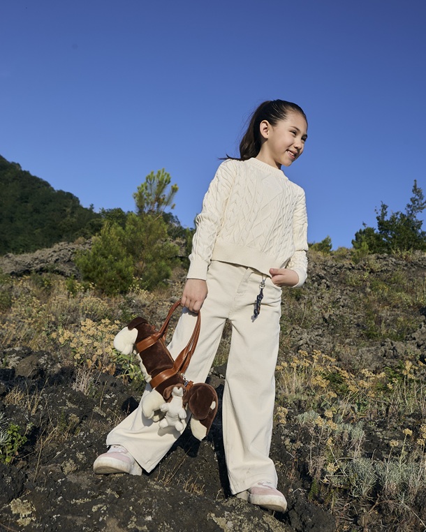 A smiling little girl outdoors on rocky ground with vegetation, dressed in a light cable-knit sweater, matching trousers, and athletic shoes, while holding a brown dachshund-shaped bag.
