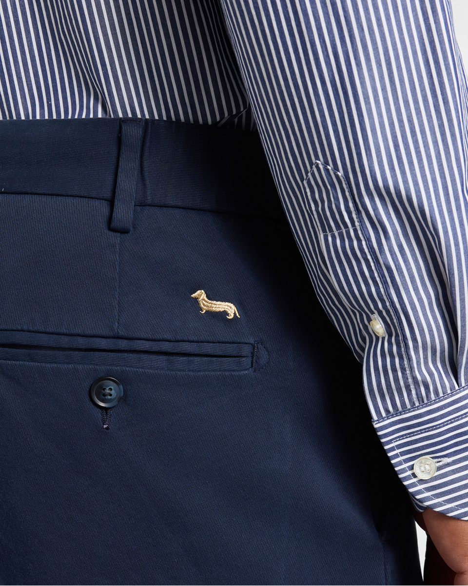 Close-up of dark blue trousers featuring an embroidered gold dachshund logo above the back pocket, paired with a blue and white striped shirt.