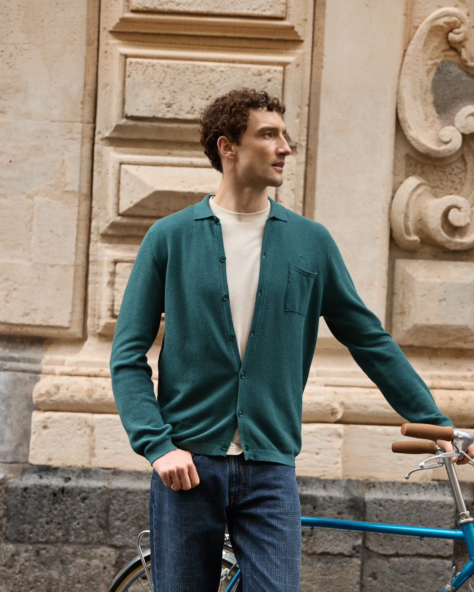Uomo in piedi accanto a una bicicletta d'epoca azzurra davanti a un palazzo storico in pietra, che indossa un cardigan in maglia verde ottanio con colletto, taschino sul petto e bottoni, portato aperto su una t-shirt panna e abbinato a jeans blu scuro.
