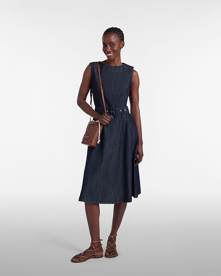 Promotional image focused on the dresses category: a smiling model poses facing forward against a light gray gradient background. she wears a sleeveless dark denim blue midi dress featuring a boat neckline and a slightly flared skirt, cinched at the waist with a matching belted buckle. the look is completed with a brown leather crossbody bucket bag and flat leather lace-up sandals.