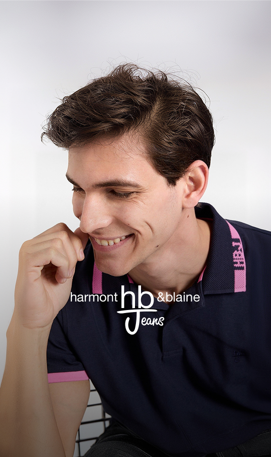 Immagine promozionale per la collezione Autunno/Inverno 2026 raffigurante un uomo e una donna che camminano sorridenti affiancati lungo un porticato storico in pietra; l'uomo indossa un cardigan a quadri verde e blu scuro su t-shirt bianca, jeans e cappellino con visiera verde oliva, mentre la donna indossa uno chemisier lungo in denim blu scuro cinturato in vita, stivali alti in camoscio marrone e porta una borsa a spalla nera.