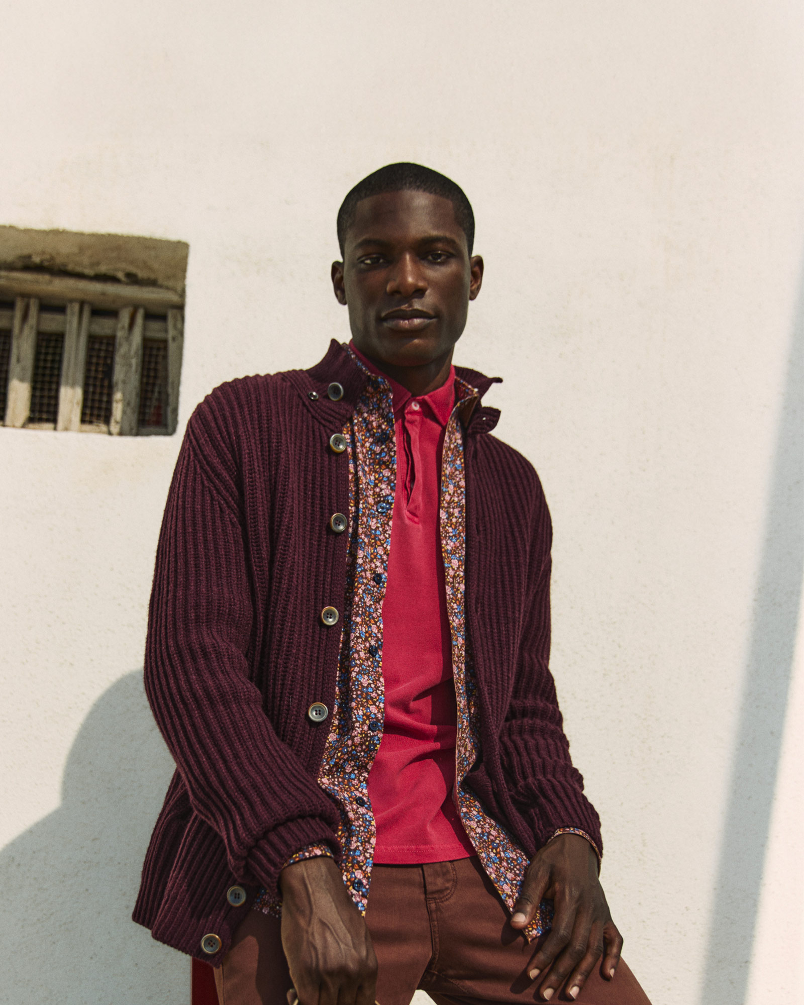 Hombre con cárdigan burdeos y camisa estampada frente a una pared clara.