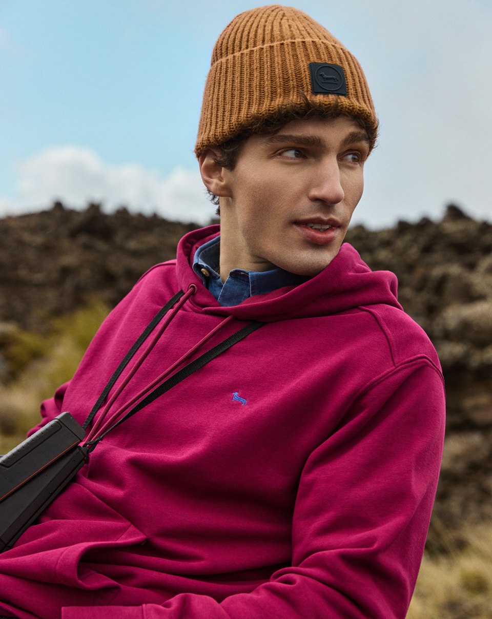 Close-up of a male model wearing a magenta hooded sweatshirt, a denim shirt, and an ochre knit beanie, posing outdoors against a rocky background.