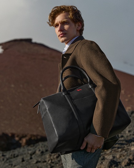 Male model wearing a brown coat and white shirt holding a large black leather travel bag under his arm, against a rocky volcanic landscape background.