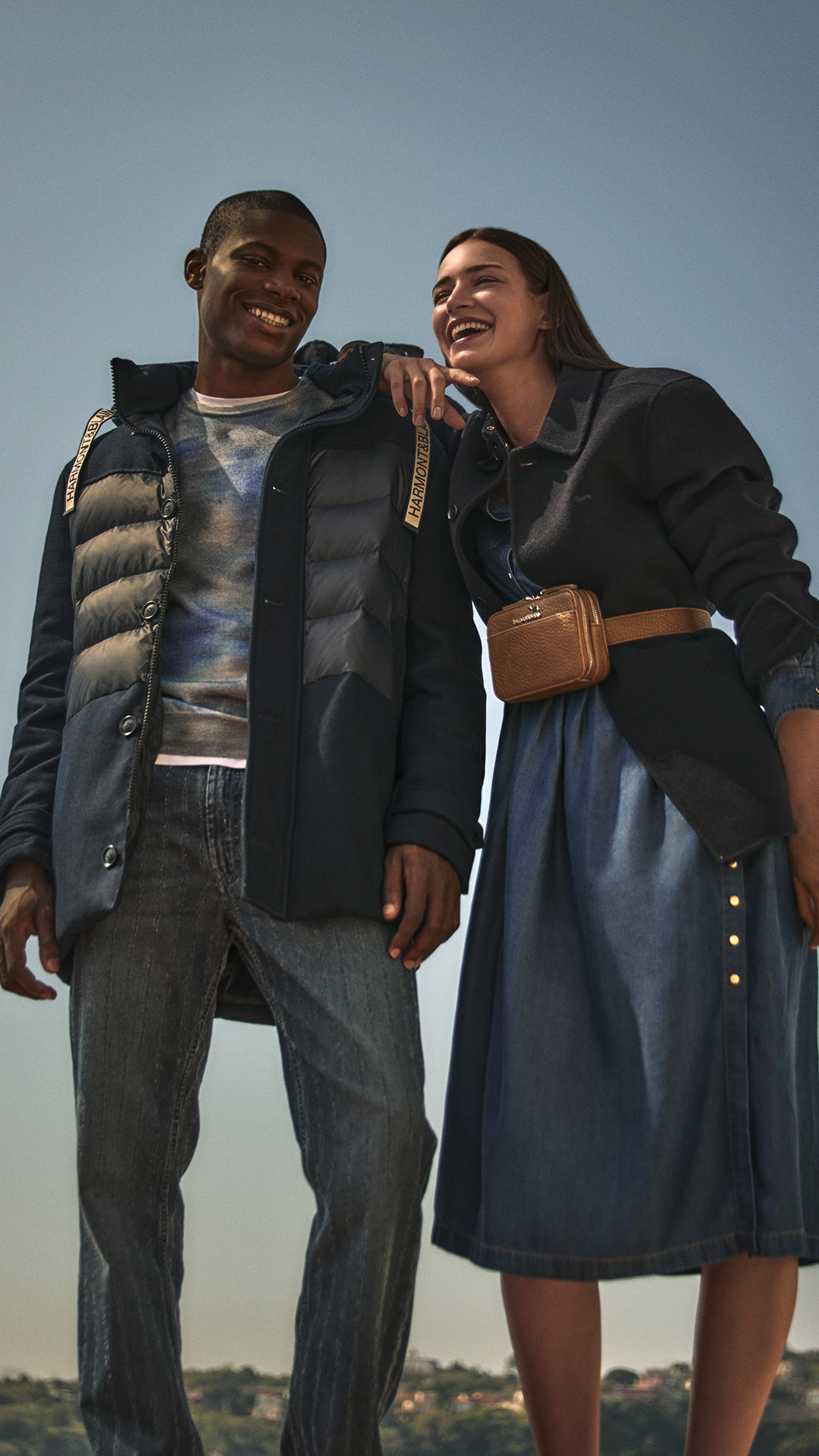 Coppia che sorride all’aperto: lui indossa un piumino blu e jeans, lei un abito in denim e giacca blu con borsa in vita.