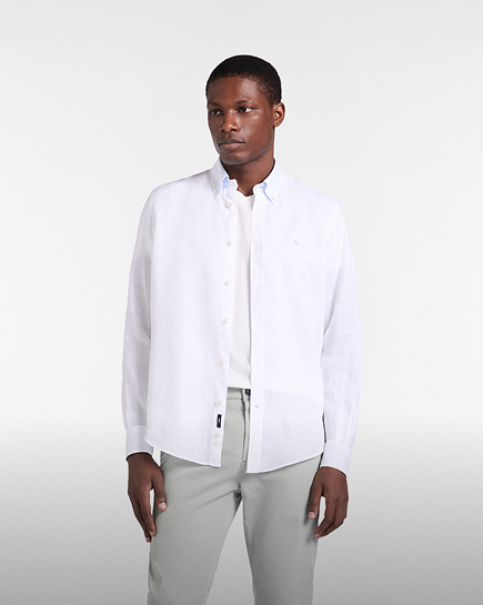 Promotional image focused on the shirts category: A model poses against a light gray background wearing a white linen long-sleeved shirt, unbuttoned at the front and worn over a white t-shirt. The shirt features a button-down collar and the dachshund logo embroidered on the chest in a matching light tone. The look is completed with light gray trousers.