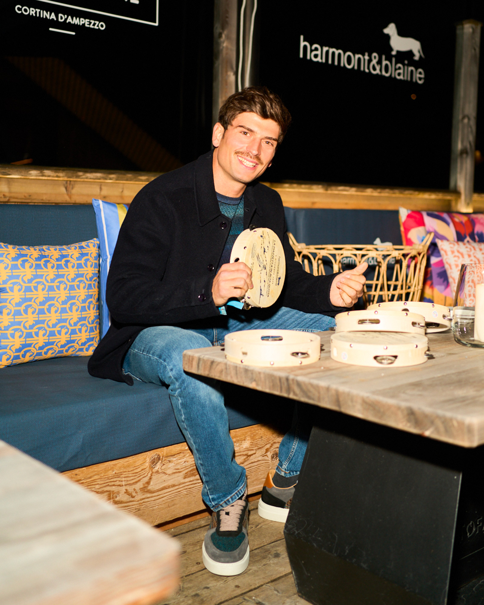 Uomo seduto con tamburello, indossa giacca blu scuro, maglia rigata e jeans.