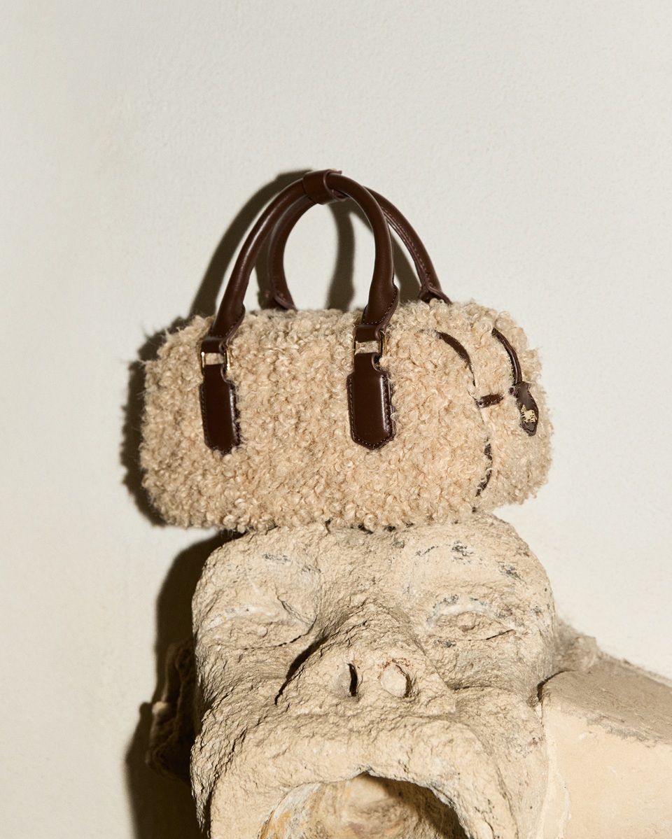 Beige shearling bowling handbag with dark brown leather handles and details, resting on top of a stone sculpture.