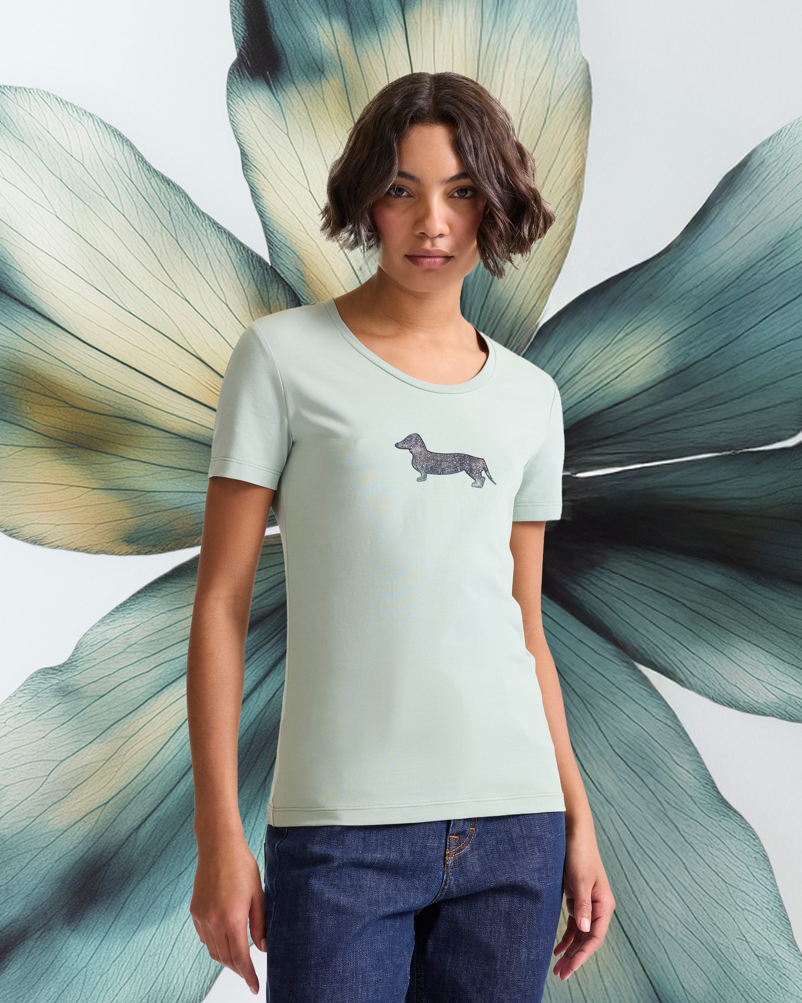 Woman wearing a light green t-shirt with dachshund logo in front of a large flower.