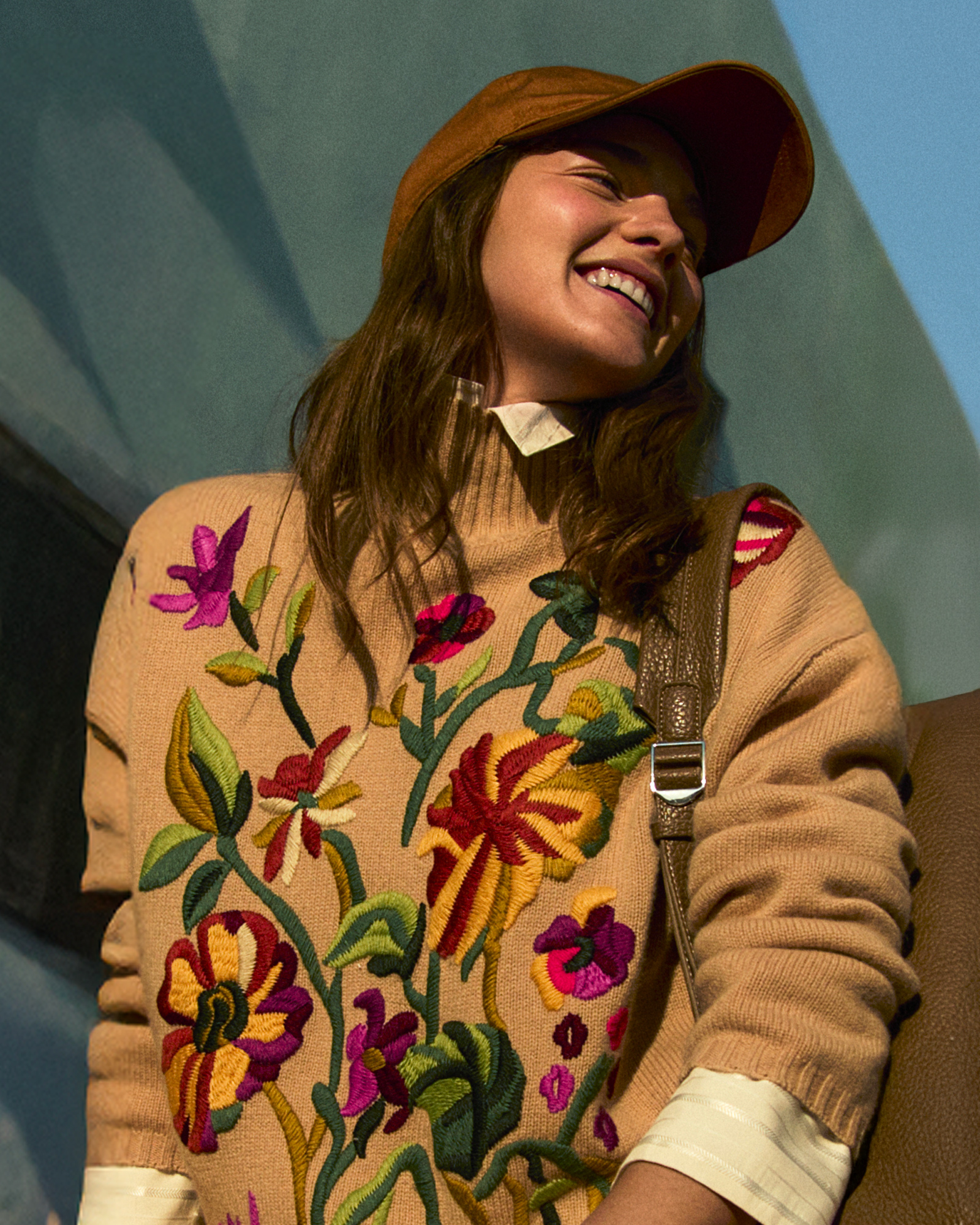 Smiling woman wearing a beige sweater embroidered with colorful flowers and a brown cap.