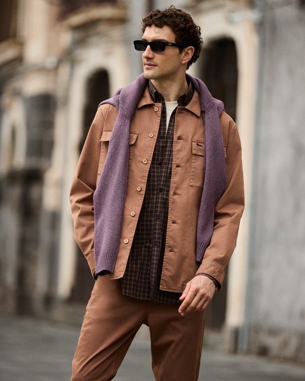 The promotional image for the Men's Outerwear Focus features a model in an urban setting wearing a tobacco-colored overshirt jacket with chest pockets, paired with matching trousers, a brown checkered shirt, and a purple sweater draped over his shoulders, accessorized with dark sunglasses.
