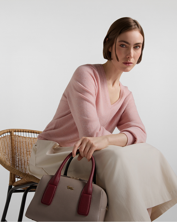 Woman seated on a wicker chair wearing a pink V-neck sweater and light-coloured skirt, holding a beige bowling bag with burgundy handles and a gold logo.