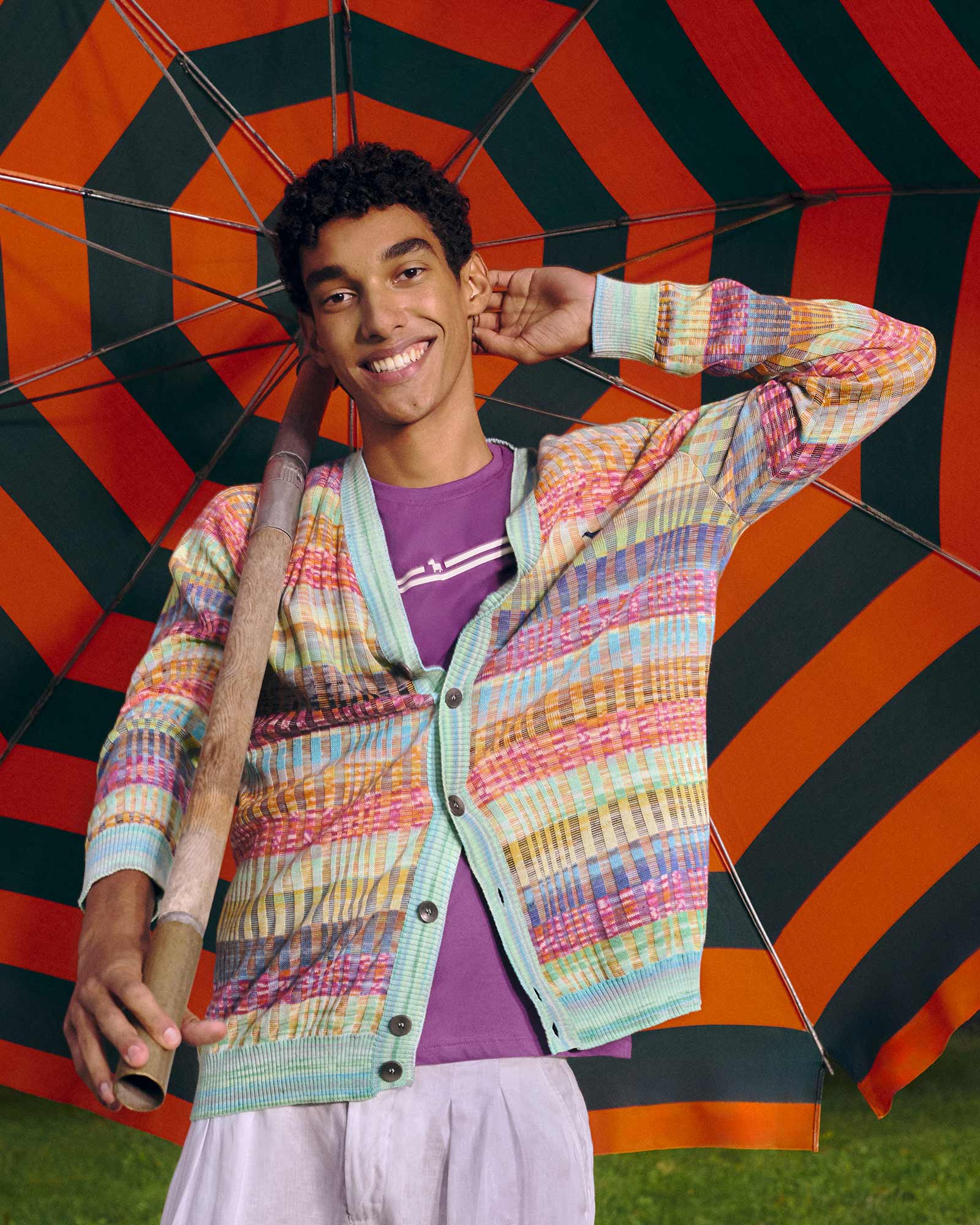 Smiling young man holding a striped beach umbrella, wearing a multicolored knit cardigan over a purple T-shirt — playful and vibrant knitwear look.