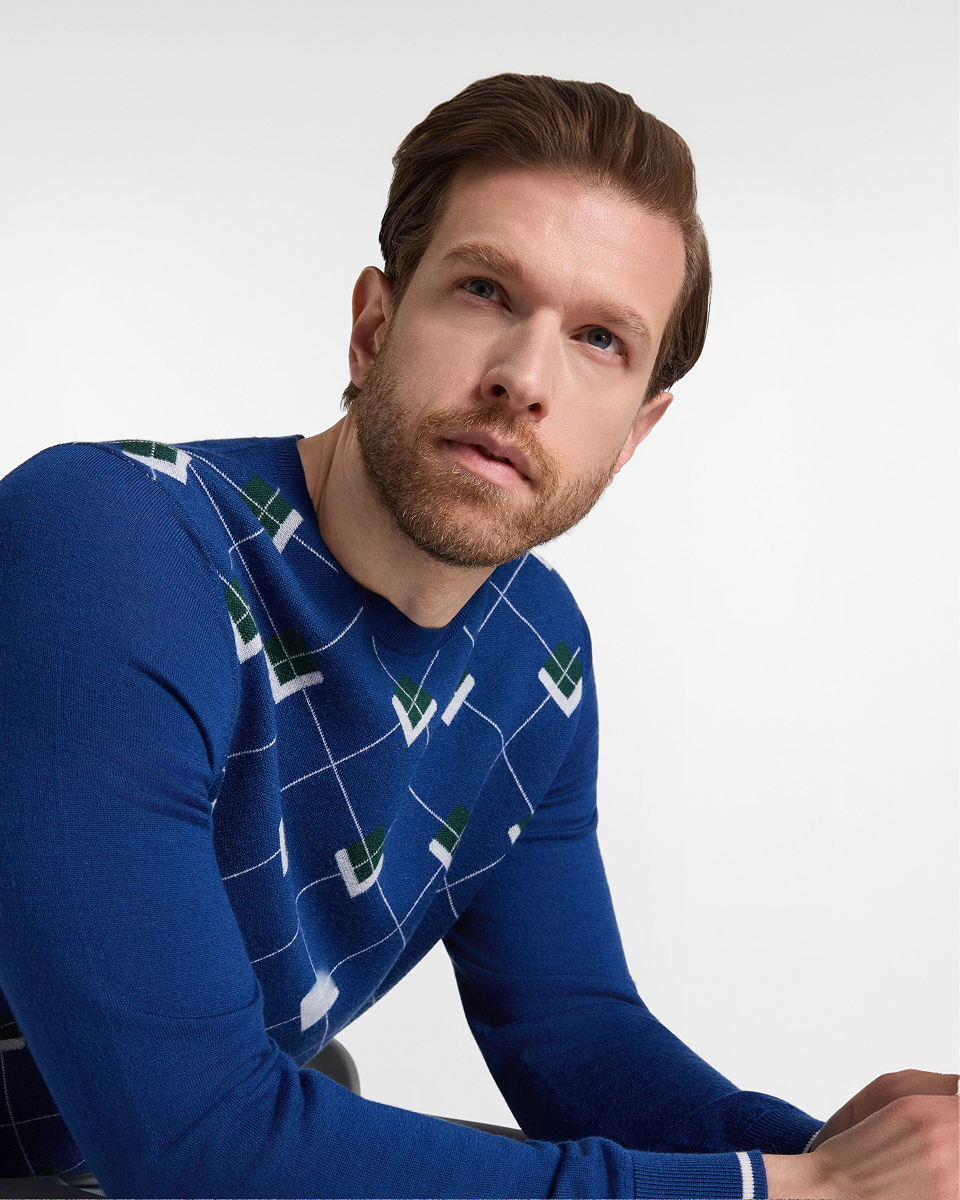 Immagine di campagna per la preview Autunno/Inverno 2026 di Harmont & Blaine, focalizzata sulla maglieria uomo. Un modello con capelli castani acconciati e una barba curata &egrave; ritratto a mezzo busto contro uno sfondo grigio chiaro, con lo sguardo rivolto verso l'alto a sinistra. Indossa un maglione girocollo blu brillante a maniche lunghe. Il capo &egrave; impreziosito da un motivo Argyle geometrico bianco su tutta la superficie, con tocchi di verde scuro all'interno dei rombi bianchi sulle spalle. Un sottile dettaglio a righe bianche orizzontali &egrave; visibile sul polsino.