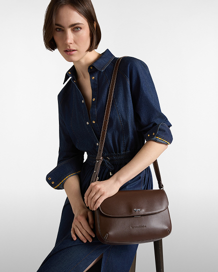 A woman with short brown hair wearing a dark blue long-sleeved denim dress with a dark brown leather crossbody bag featuring a flap closure.