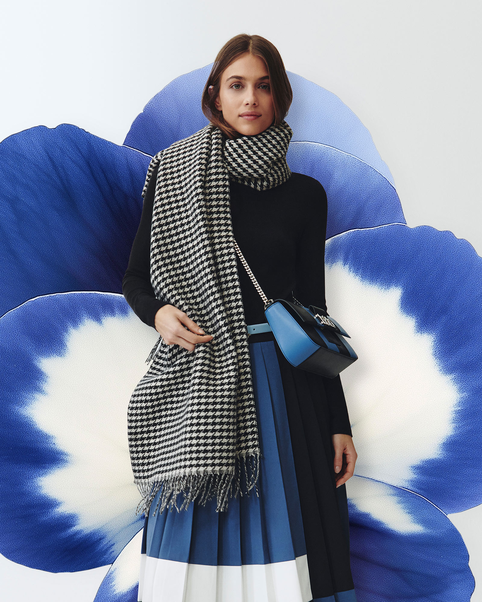 Woman wearing a houndstooth scarf, black top, blue pleated skirt and a blue shoulder bag.