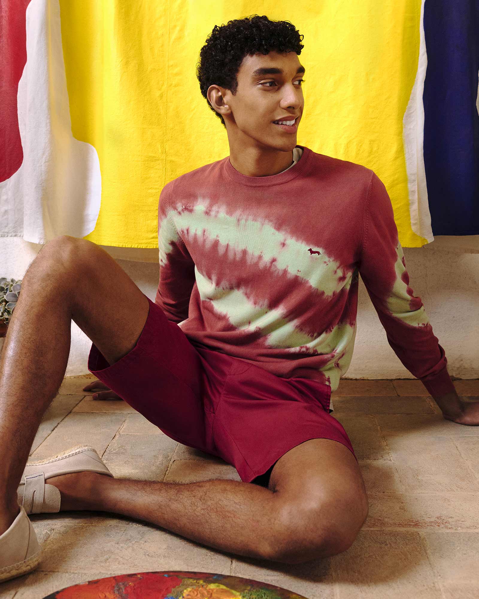 Young man sitting on the floor wearing red Bermuda shorts and a tie-dye sweatshirt, with colorful towels hanging in the background — casual and vibrant summer look.