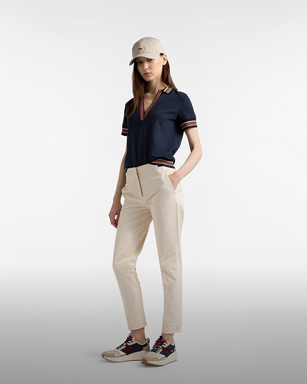 Promotional image focused on the women's trousers category: a model poses in a three-quarter view against a light gray gradient background. she wears a pair of tailored chino trousers in light beige, with a mid-rise waist and ankle-length cut. the look is paired with a dark blue polo shirt featuring multicolored striped edges, a beige baseball cap, and two-tone sneakers. the model has her left hand in her trouser pocket.