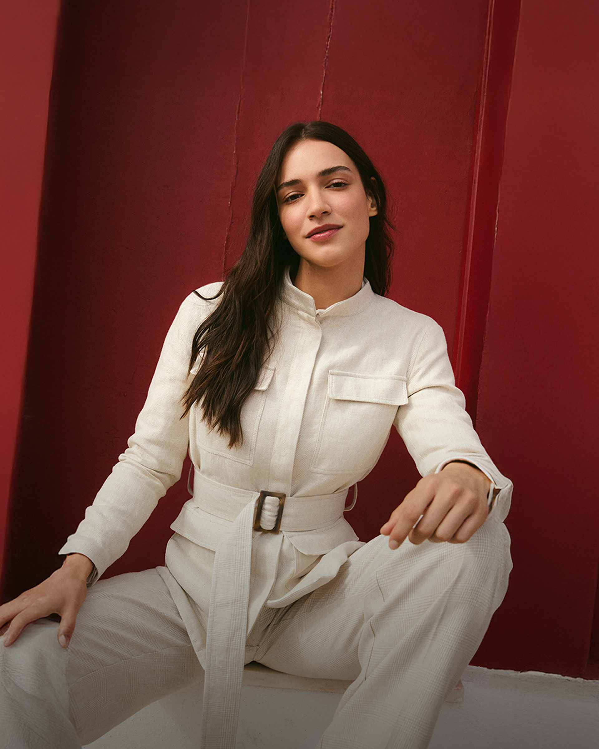 Woman in a cream-colored safari jacket with a belt, standing in front of a bright red wall. The text: made in Italy for her