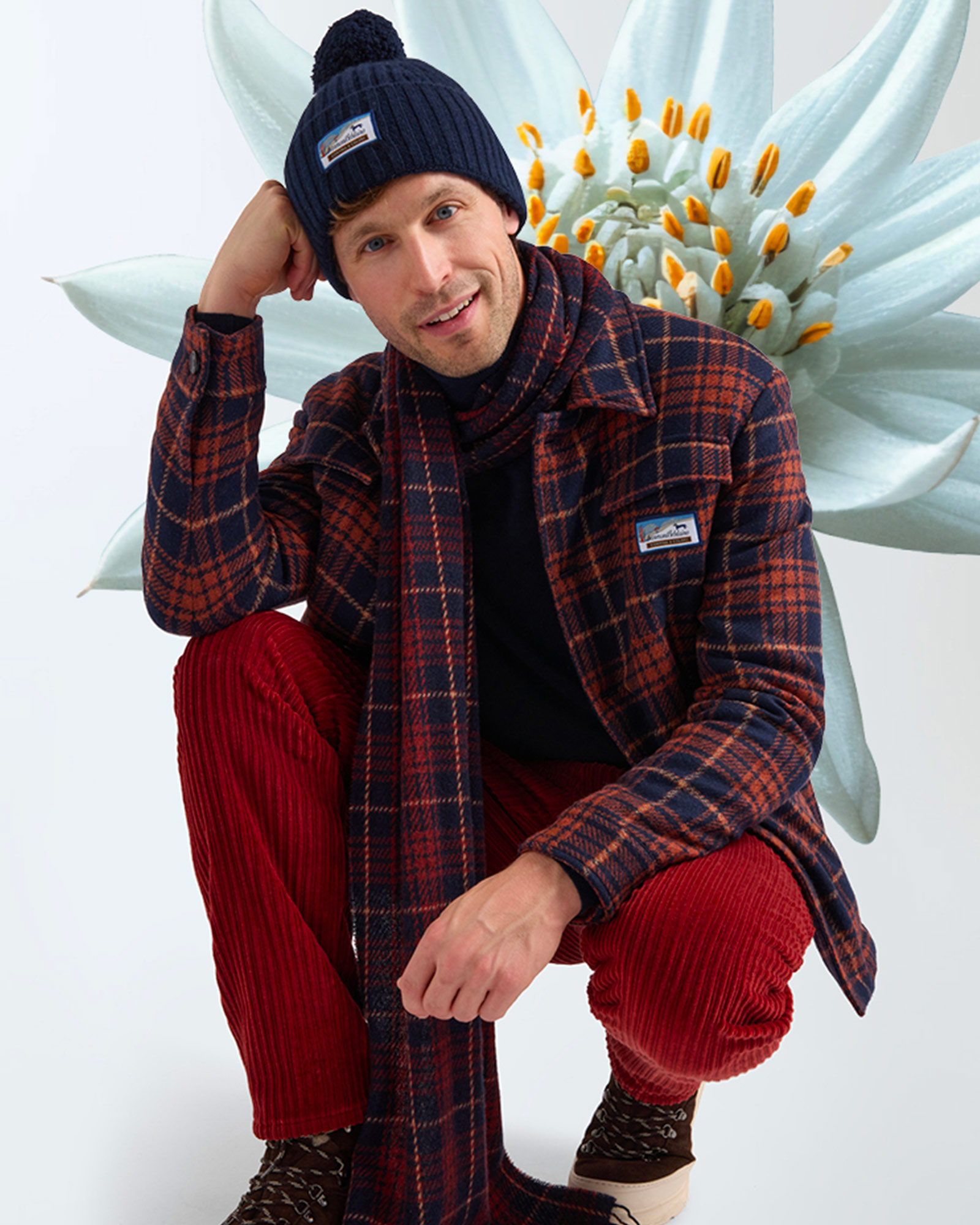 Man crouching wearing navy beanie, blue and red checked overshirt and red corduroy trousers.