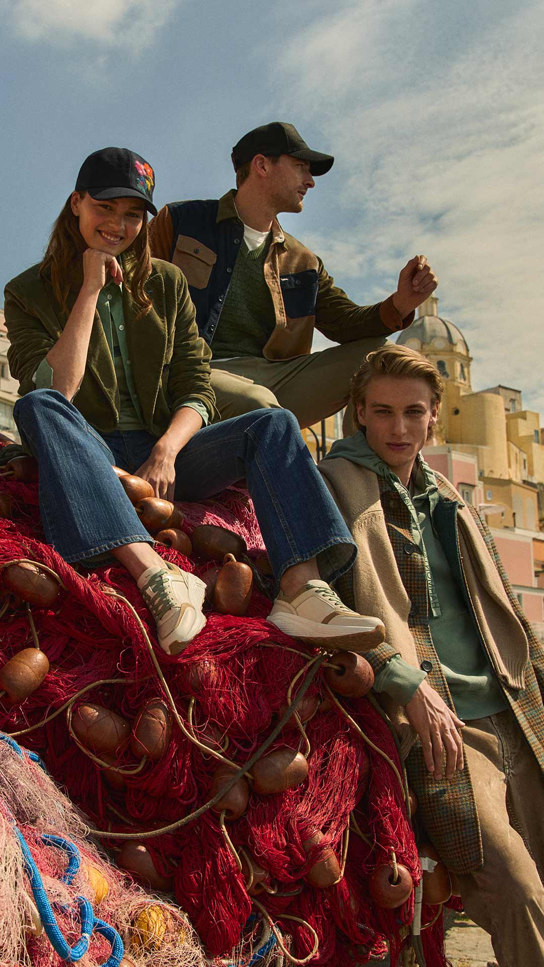 Three young models sitting on colorful fishing nets in a picturesque Mediterranean village, wearing casual layered outfits with a relaxed, coastal vibe.