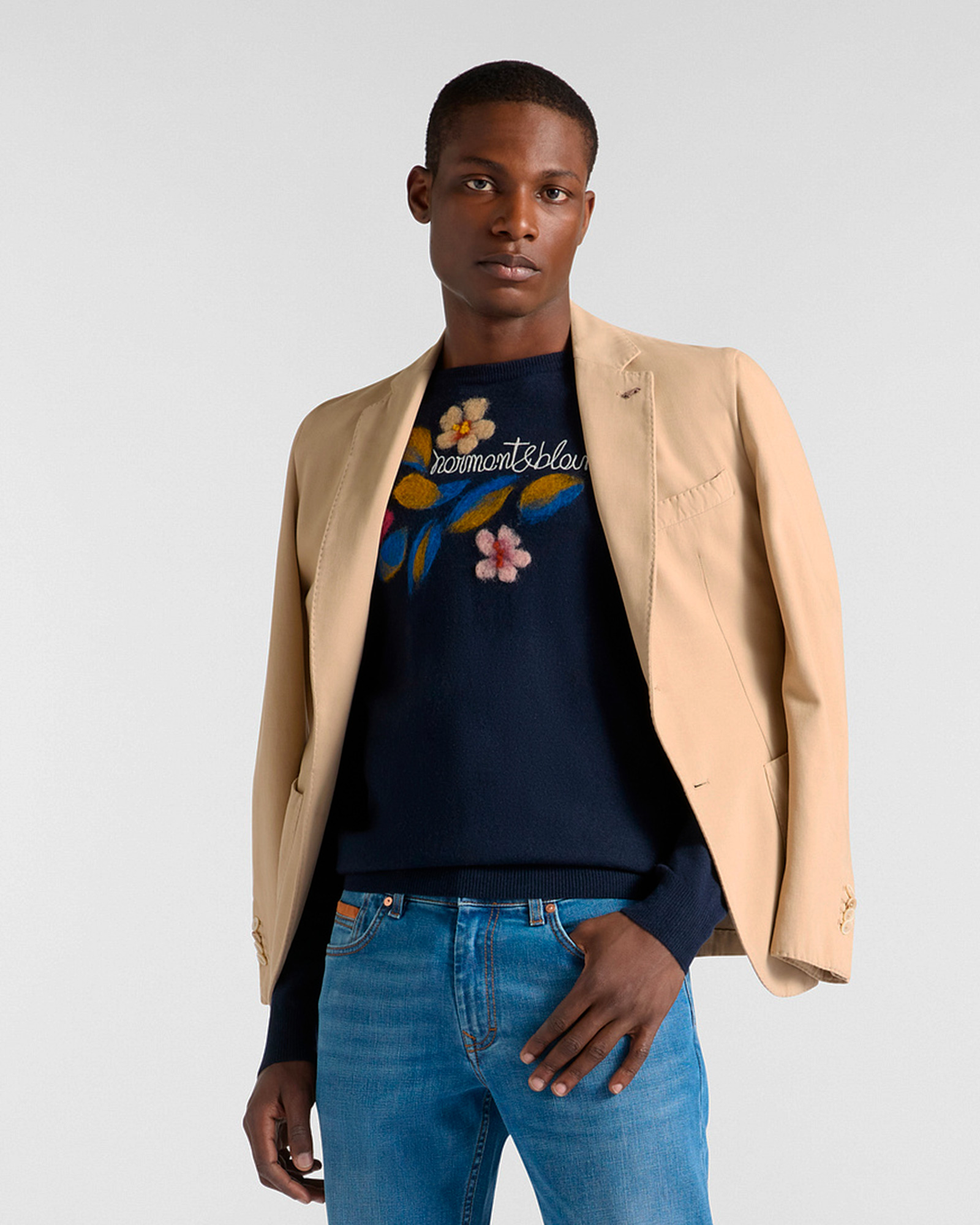 Man in beige blazer over navy sweater with floral embroidery and jeans