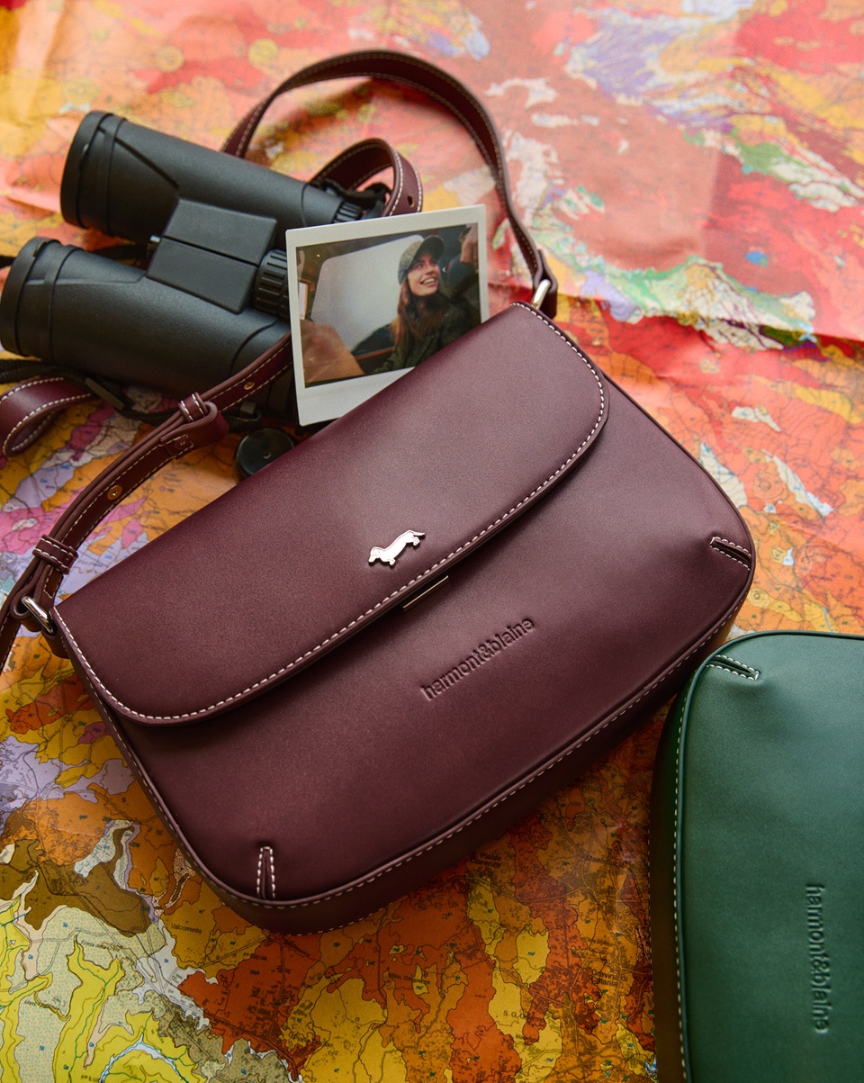 Burgundy leather crossbody bag with a metal logo resting on a geographical map, alongside black binoculars, an instant photo, and a green bag.