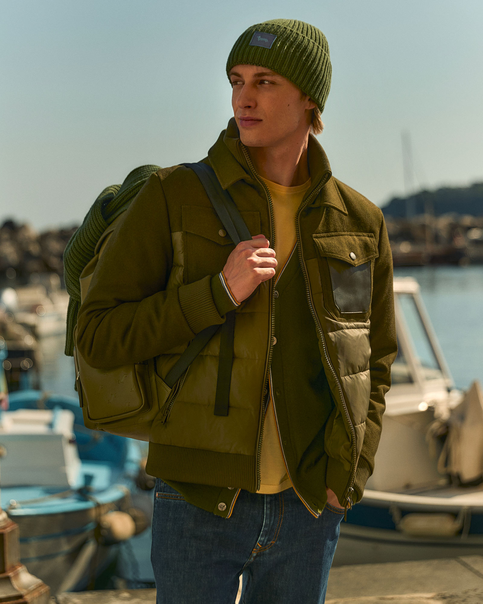 Man wearing a layered green jacket, green beanie, and backpack near the harbor.
