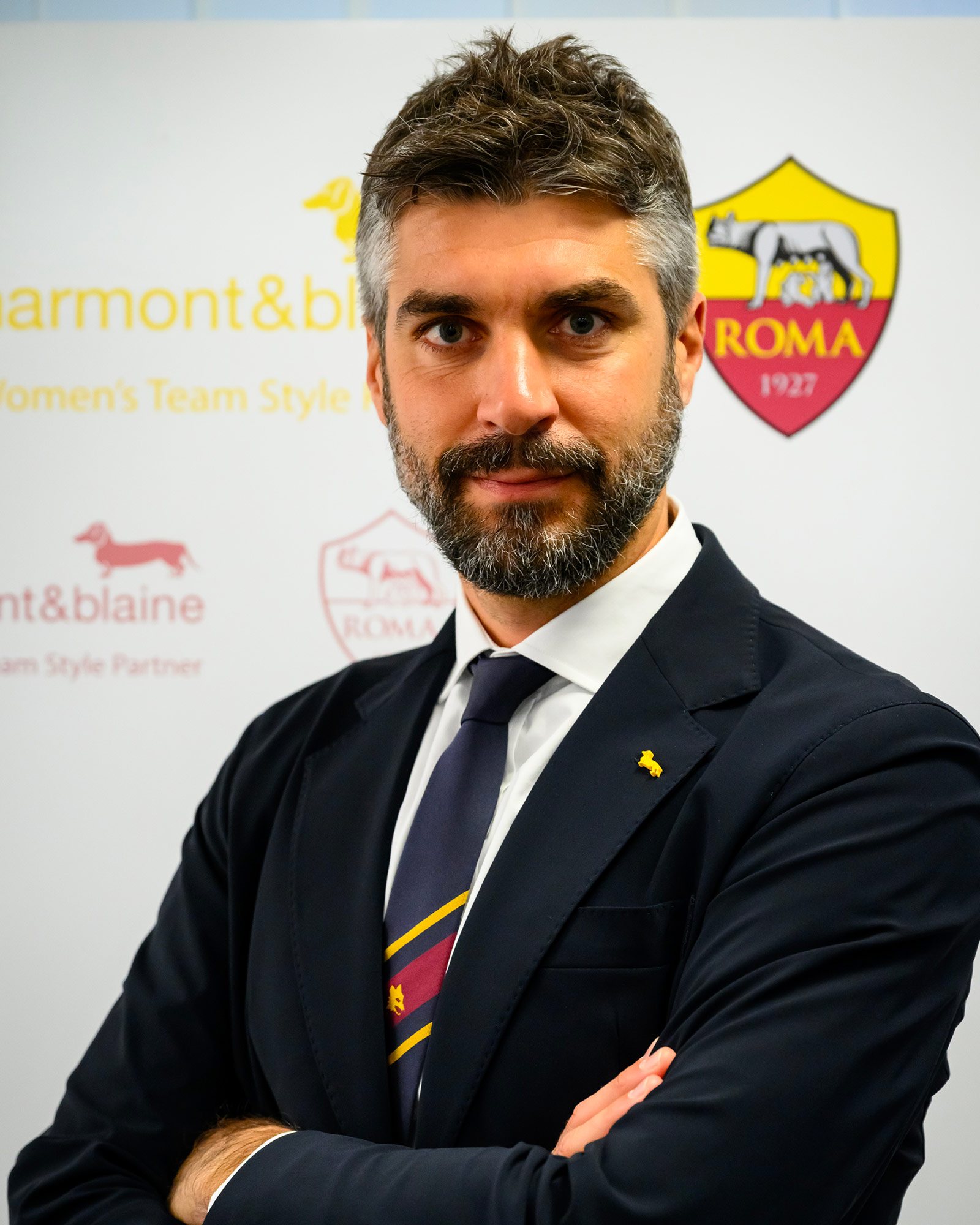 Uomo in abito blu navy con cravatta a righe e spilla gialla.