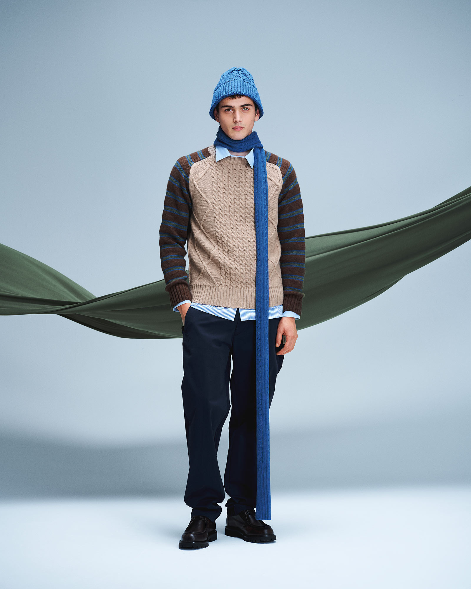 Man wearing beige cable knit sweater, dark trousers and blue beanie with matching scarf