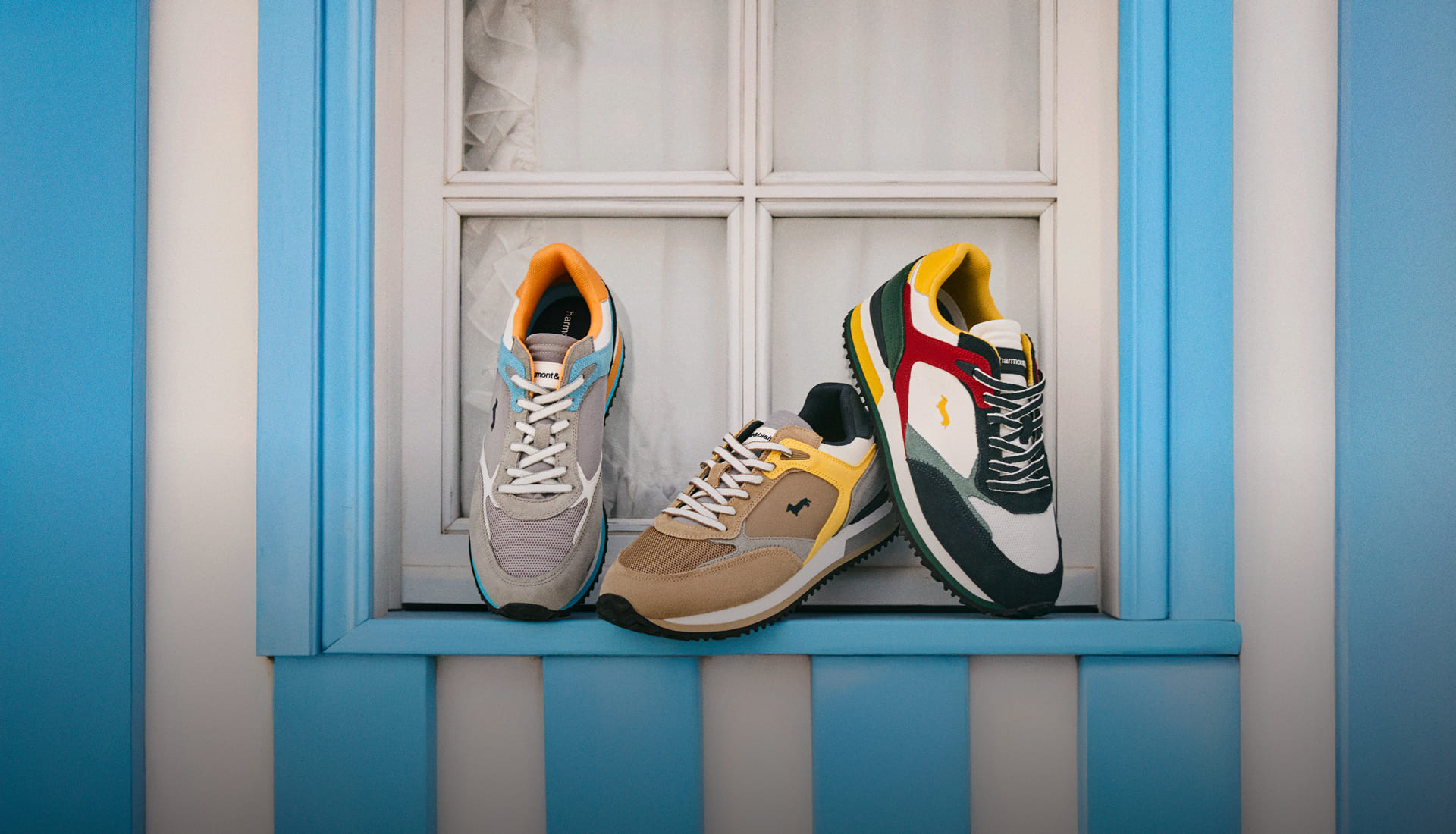Three colorful sneakers (in grey/orange, beige/brown, and green/red/yellow tones) are displayed in a row on the light blue sill of a white window.