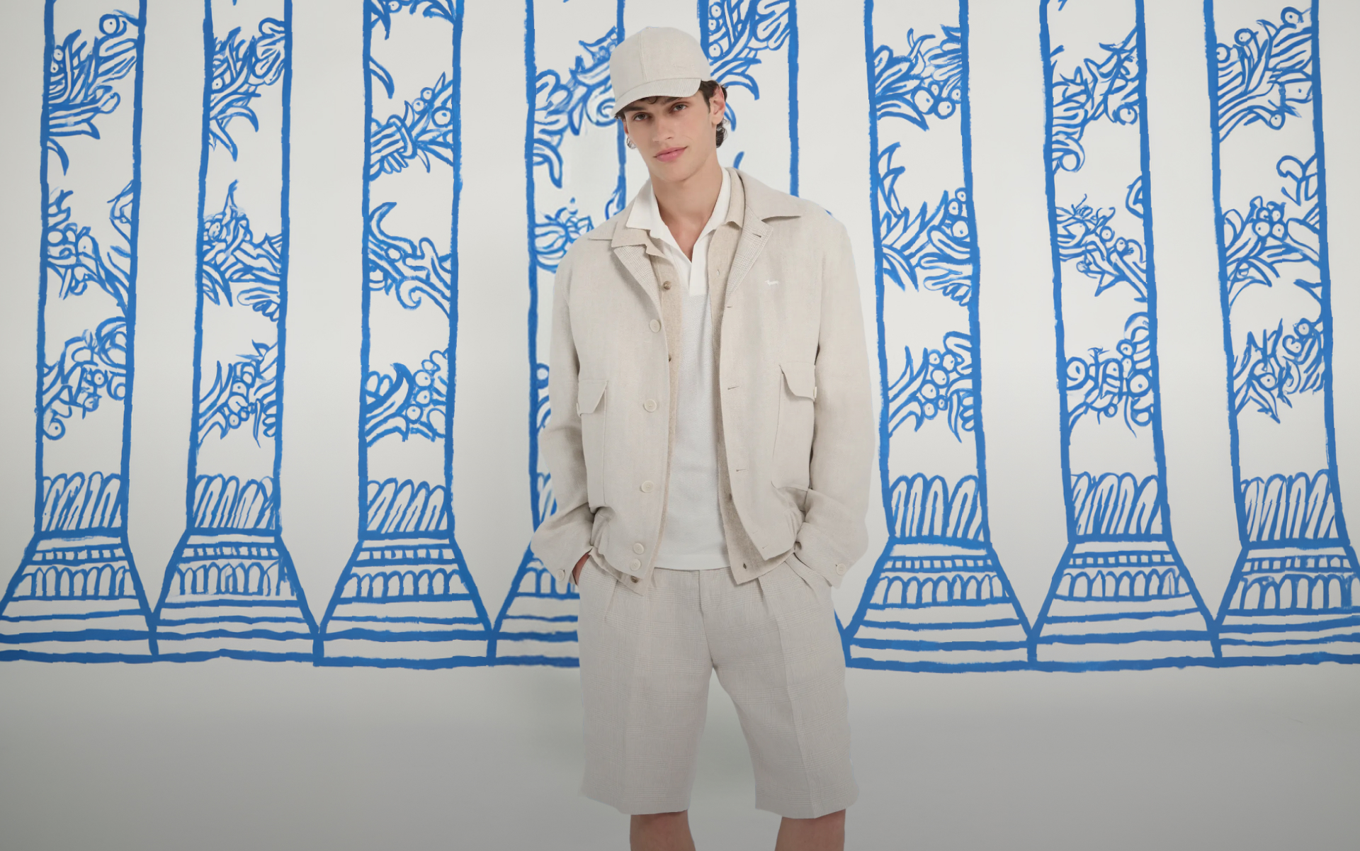 Linen Collection Banner: A model in a total-white outfit, including a hat and jacket, against a yellow background decorated with blue drawings of umbrellas and palm trees. Text: Linen Collection. Freshness and Freedom. Link to the Linen Collection.