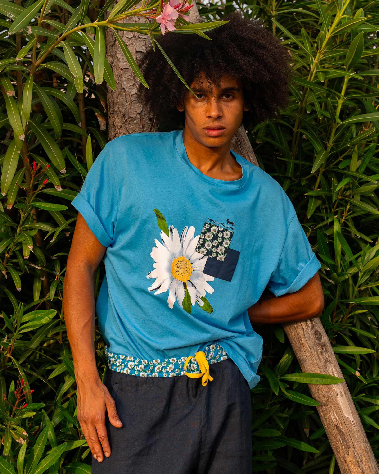 Young man wearing a light blue T-shirt with a large daisy print and floral patchwork detail, standing in front of lush greenery — bold and fresh summer look.