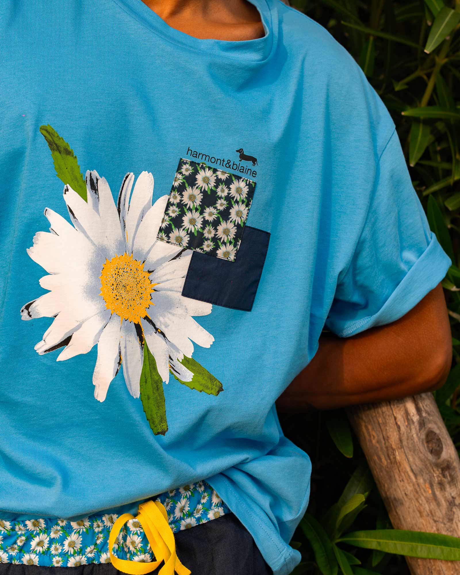 Close-up of a bright blue T-shirt with a large daisy print and floral patchwork details, featuring the Harmont & Blaine logo.