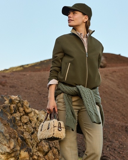 Female model wearing an olive green jacket, matching cap, patterned trousers, and a sweater tied around her waist, holding a small faux-shearling handbag against a rocky volcanic background.