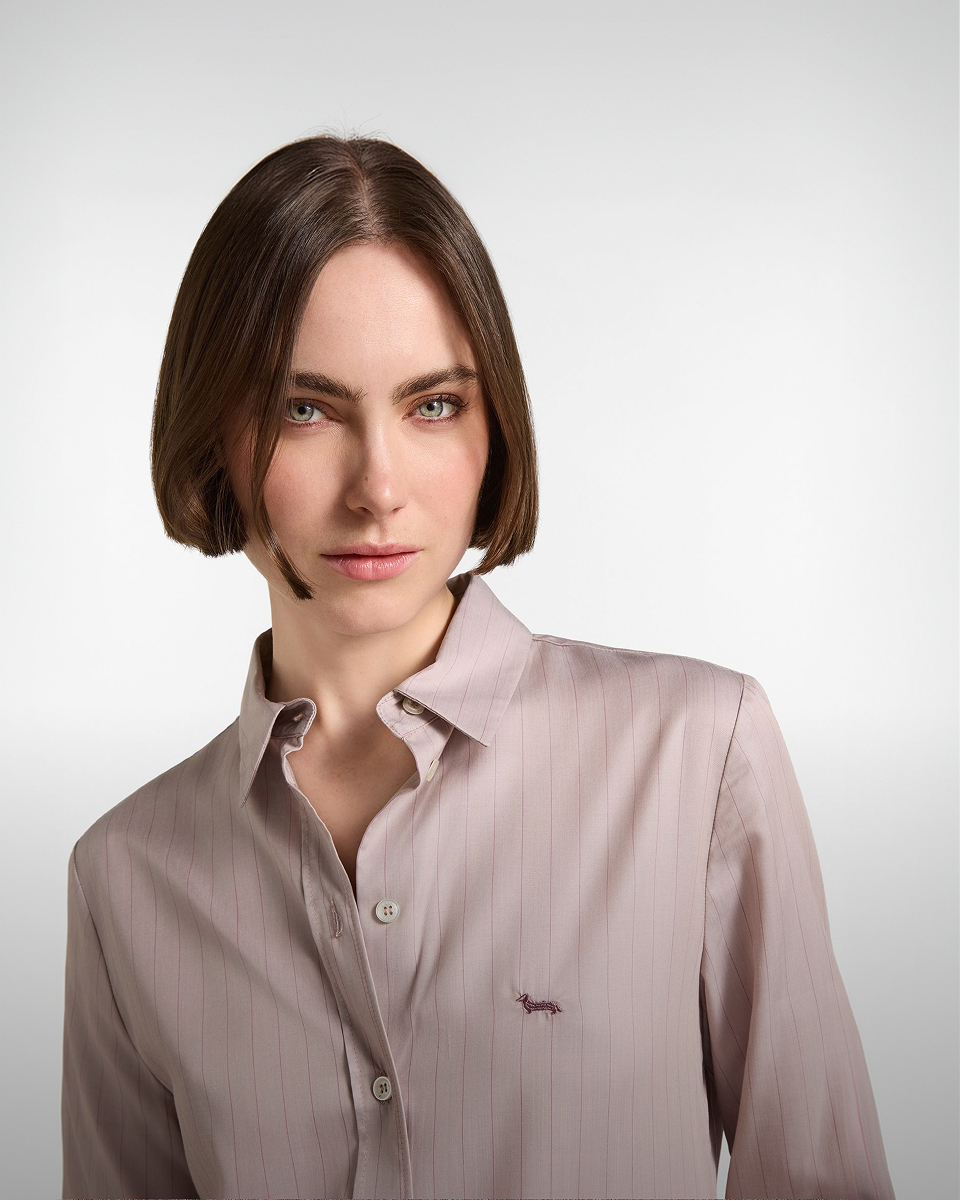 Woman with a bob haircut wearing a dusty pink pinstriped shirt featuring an embroidered dachshund logo on the chest.