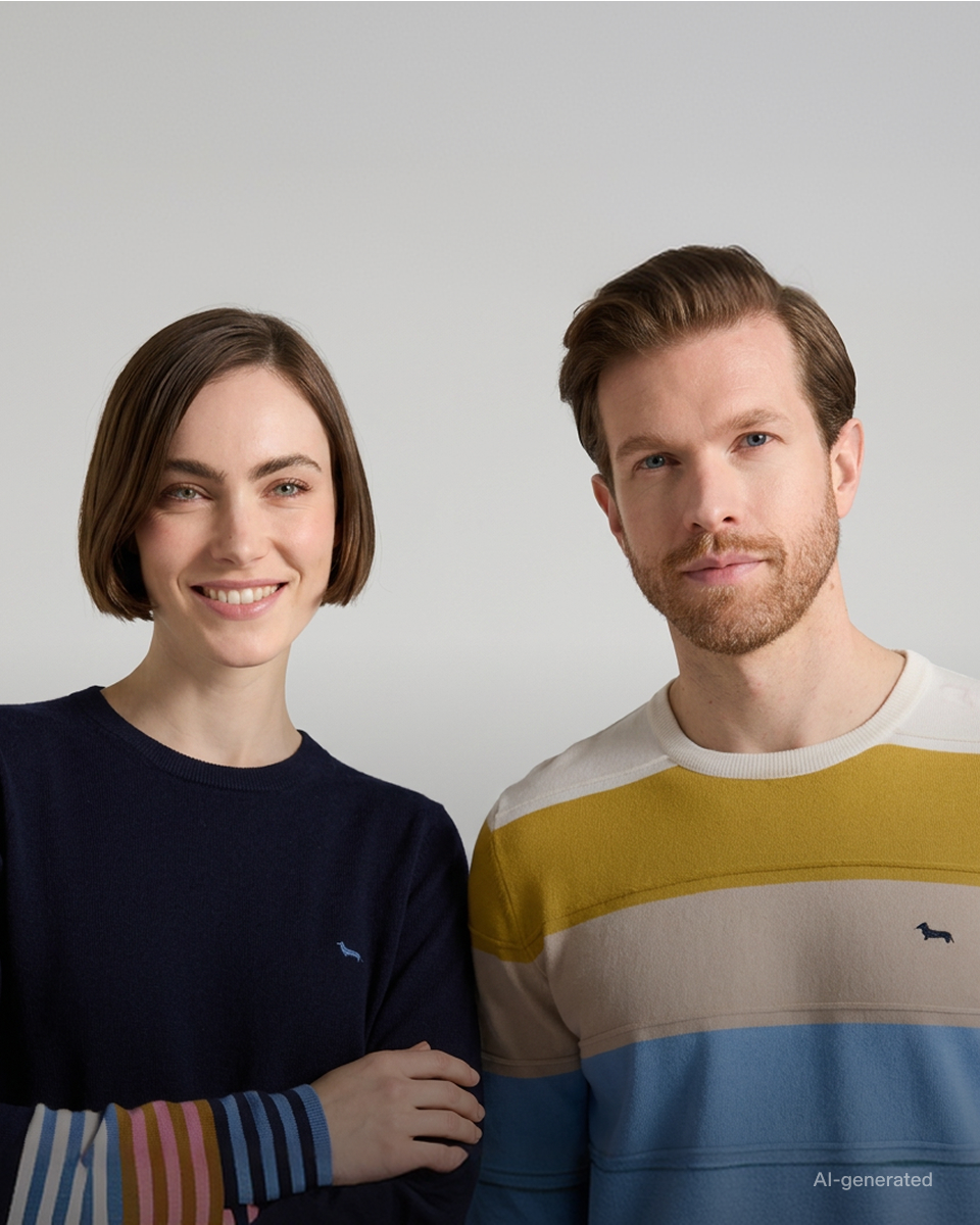 AI-generated campaign image for the FW26 collection, themed "Stripes for Her and for Him". The photo portrays a half-length shot of a woman and a man side by side against a neutral gray background. The smiling woman on the left, with a short brown bob, wears a dark blue crewneck sweater featuring multicolored striped cuffs in shades of pink, light blue, and ochre, with a small embroidered logo on the chest. The man on the right, with a beard and brown hair, sports a crewneck sweater with wide horizontal stripes in ochre, cream, and light blue, with the dachshund logo embroidered at the center of the chest.