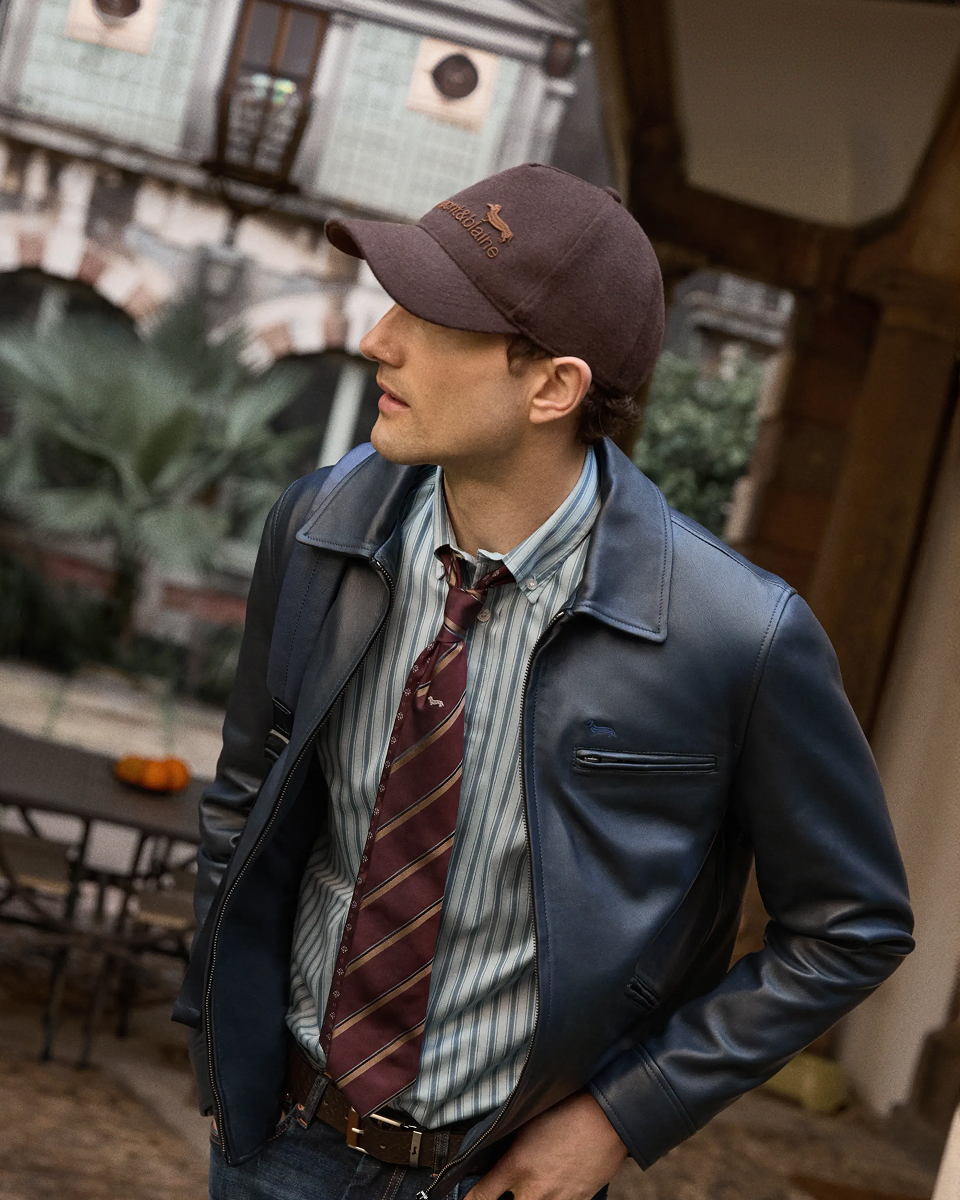 Uomo inquadrato a mezza figura all'aperto che indossa un giubbotto in pelle blu scuro con colletto classico e tasca sul petto, abbinato a una camicia a righe verticali azzurre, cravatta bordeaux a righe trasversali, jeans e un cappellino in panno marrone con logo ricamato.