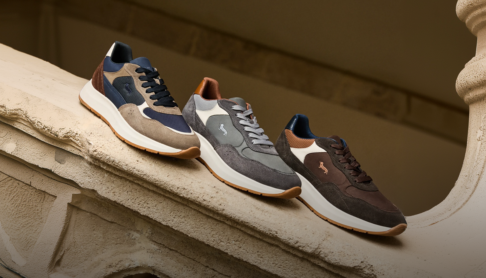 Tre paia di sneaker da uomo della collezione Autunno/Inverno 2026 disposte in fila su una balaustra in pietra decorata, realizzate in camoscio e tessuto tecnico multi-materiale nei toni del blu, grigio, marrone e beige, tutte caratterizzate dal logo del bassotto ricamato sul lato e da una suola spessa bianca con battistrada in gomma.