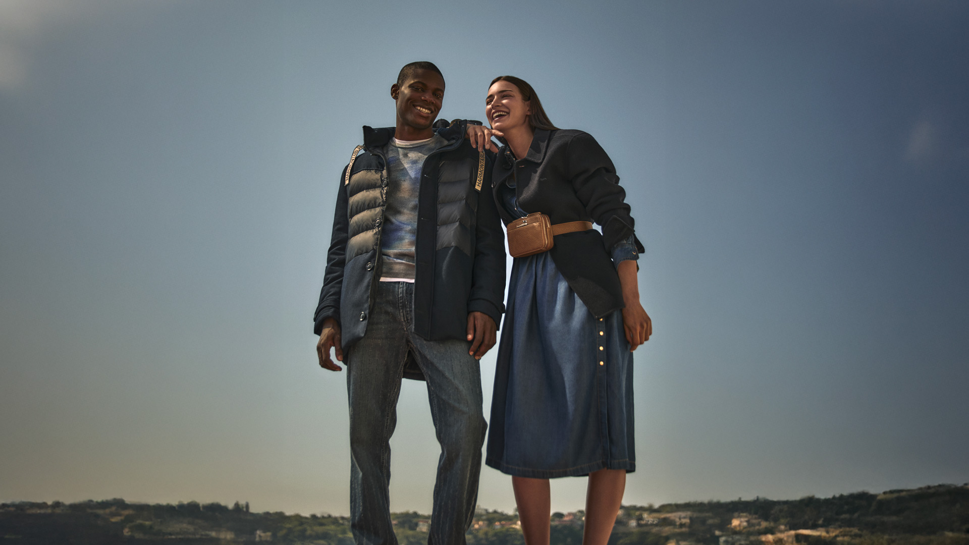 Coppia che sorride all’aperto: lui indossa un piumino blu e jeans, lei un abito in denim e giacca blu con borsa in vita.
