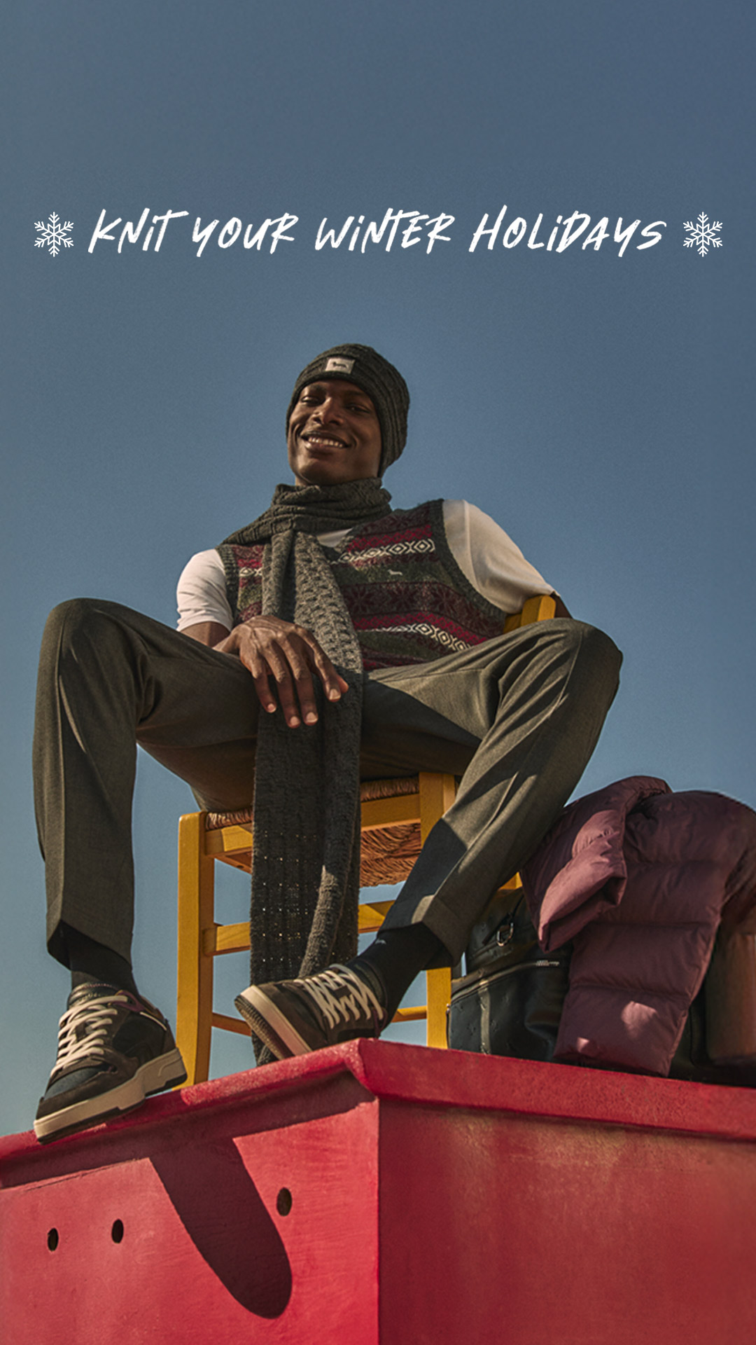 Man sitting outdoors wearing a knitted vest, grey scarf, beanie and tailored trousers.