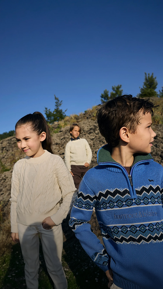 Three children are wearing Harmont&Blaine knitwear items&mdash;in the foreground, a girl in a light cable-knit sweater and a boy in a blue zip-up sweater with dark geometric patterns, while a third child in the background wears a light pullover&mdash;immersed in an outdoor natural setting under a clear blue sky.
