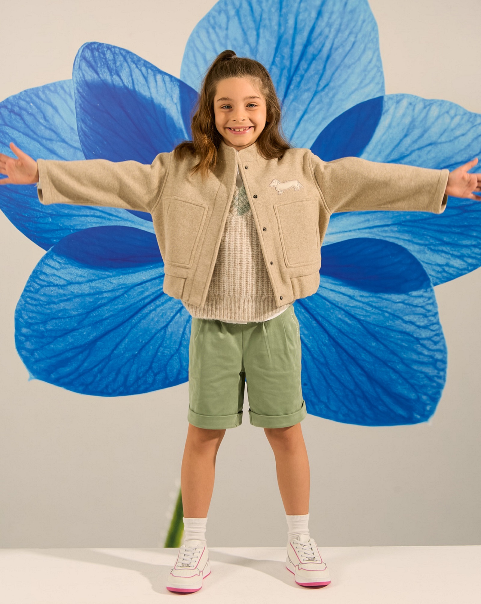 Bambina con giacca beige, maglione chiaro, bermuda verdi e sneakers bianche