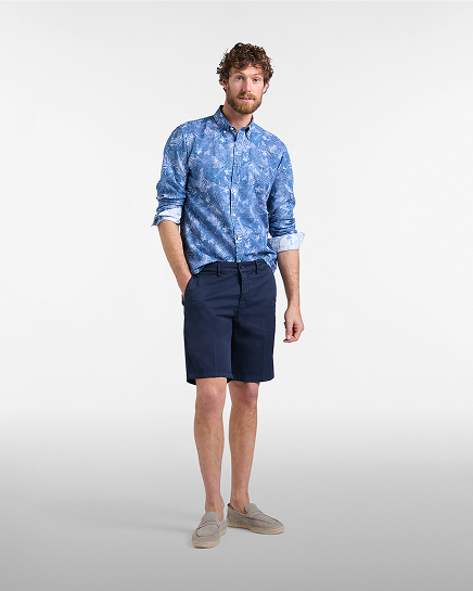 Promotional image focused on the bermuda shorts category: a man poses against a gray background wearing a pair of dark blue knee-length bermuda shorts. the look is completed with a blue long-sleeved shirt featuring a faded floral pattern and beige summer loafers.