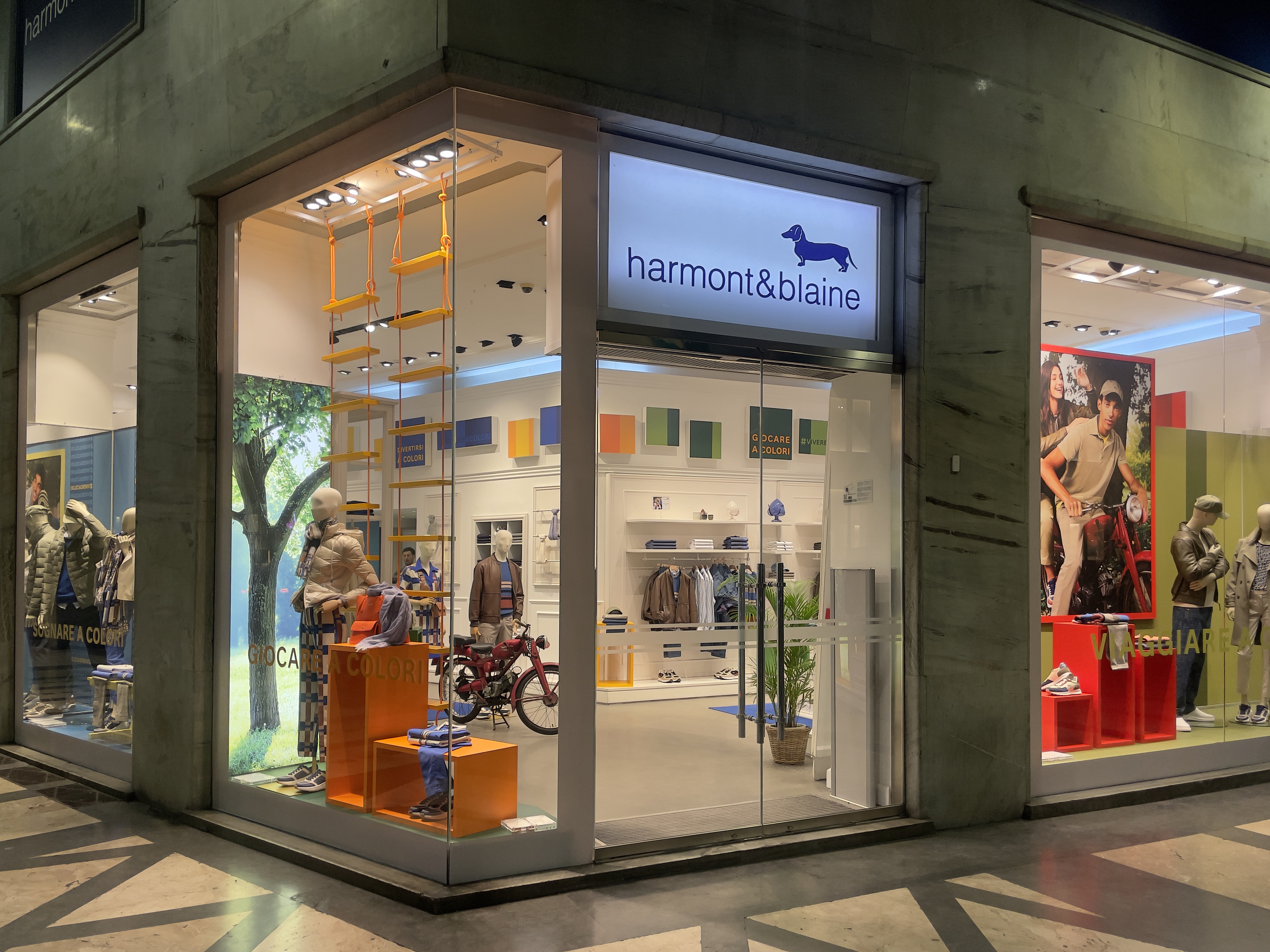 Ingresso di una boutique Harmont & Blaine con logo e due manichini vestiti in stile casual all'entrata. All'interno si vedono pareti blu decorate con quadri colorati e abbigliamento esposto. Testo in sovrimpressione: Trova la tua Boutique. Scopri la boutique pi&ugrave; vicina a te e immergiti in un&rsquo;esperienza di shopping unica.