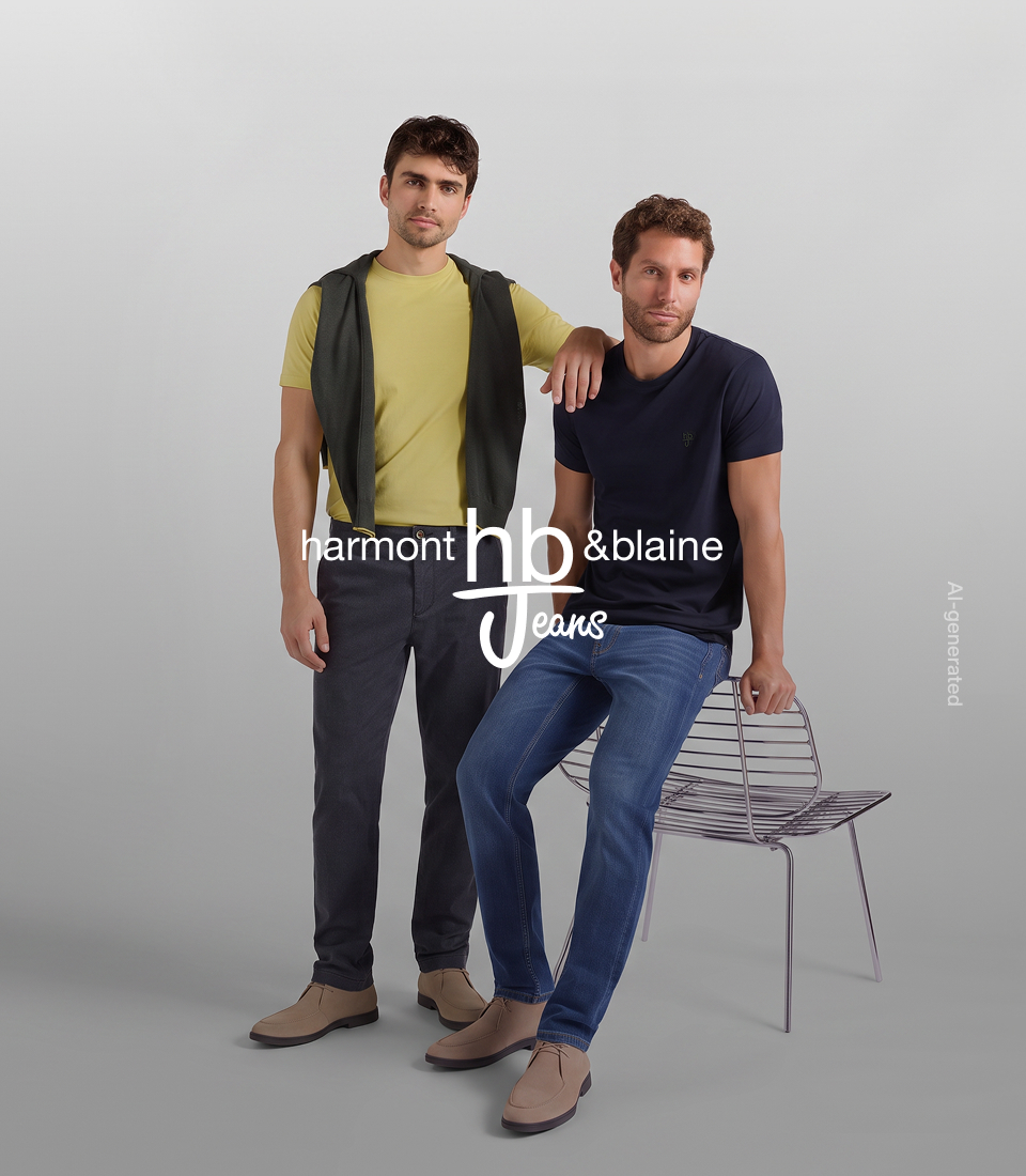 HB Jeans campaign image featuring two male models against a neutral grey background. The model on the left is standing, wearing an acid green t-shirt with a dark sweater draped over his shoulders, dark trousers, and beige suede chukka boots. The model on the right is seated on a metal chair, wearing a dark blue t-shirt, medium-wash blue jeans, and beige suede chukka boots. The white Harmont & Blaine HB Jeans logo is displayed in the center of the image. Image generated with Artificial Intelligence.